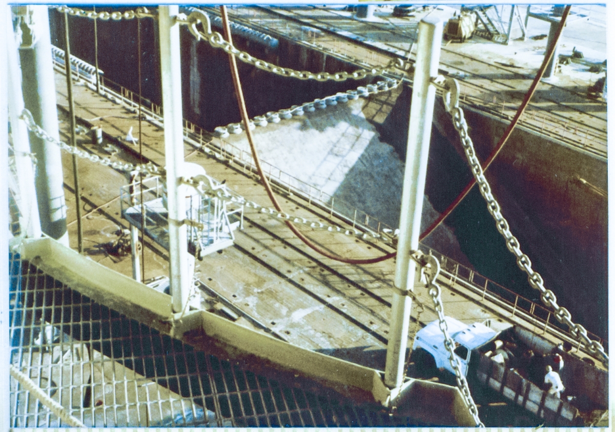 Looking directly at the Flame Deflector, sitting in the Flame Trench at Space Shuttle Launch Complex 39-B, Kennedy Space Center, Florida, from the 112’ elevation of the Rotating Service Structure, through the removable handrail which guards the curved cutout in the steel-bar grating that accommodates the orbiter’s right OMS Pod. The lower left corner of this image gives an unsettlingly-clear view of pad deck, six stories below you, through the grating panel which you are standing on.